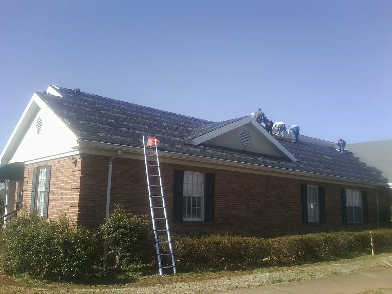 Crew tearing shingles off a brick ranch roof with a ladder set — Emergency Roof Repair work by our crew in Ellaville