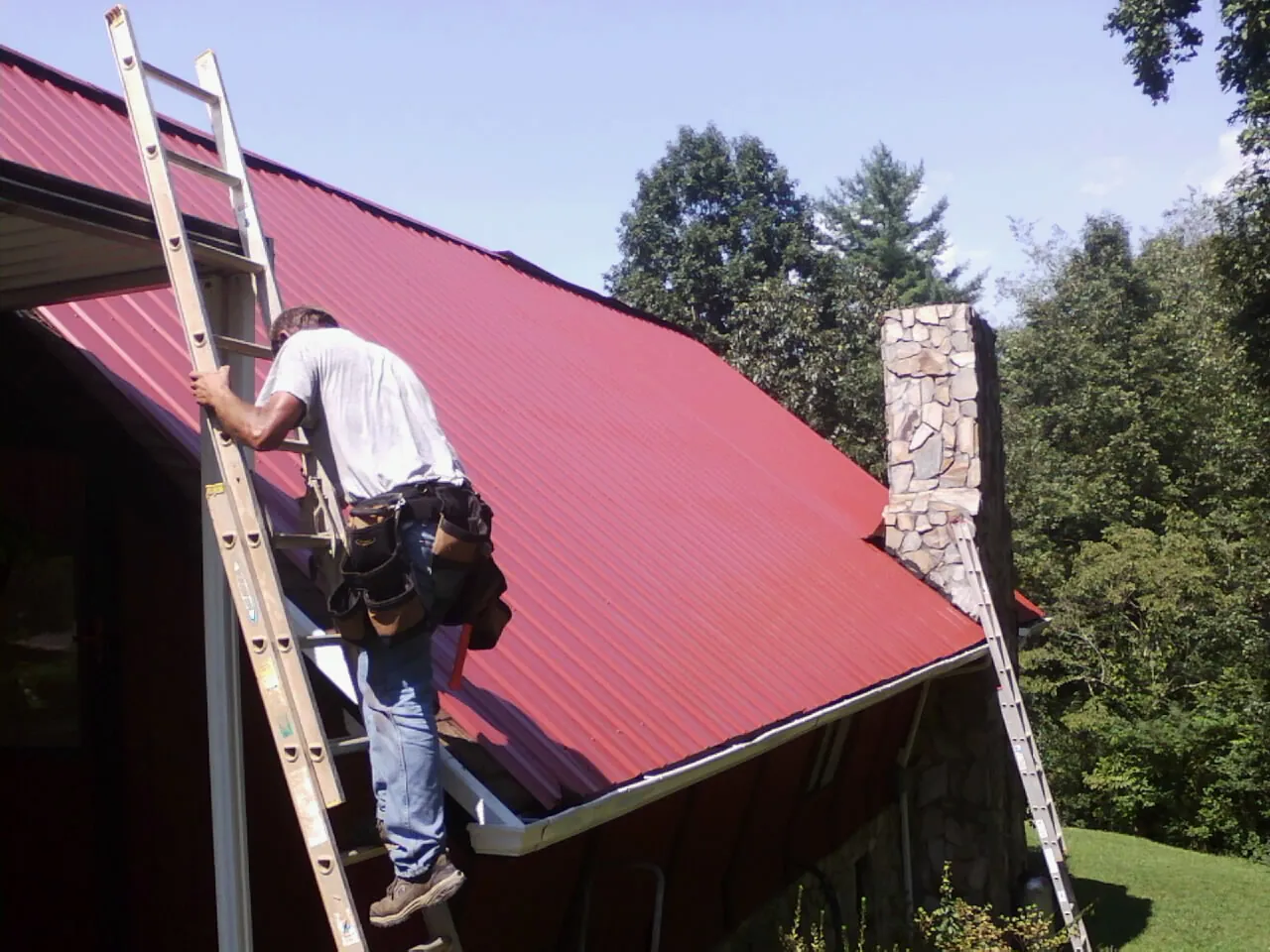 Roofer on a ladder beside a red metal roof and stone chimney — Soffit & Fascia Repair by Roofing Brothers in Ellaville